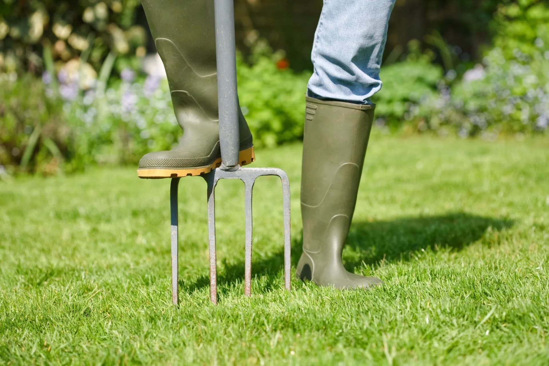 Person in rubber boots pressing a garden fork into green lawn.