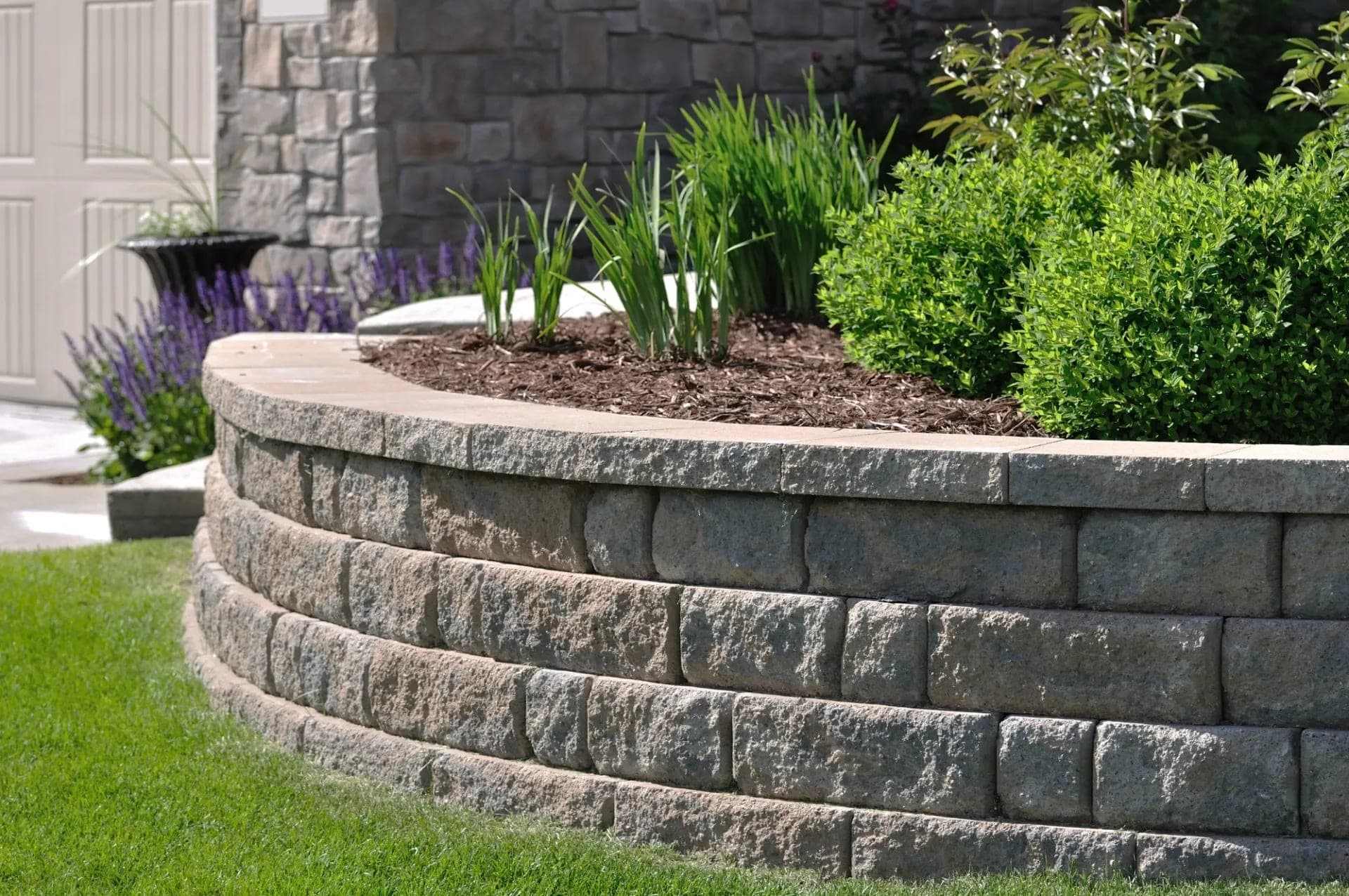 Curved block retaining wall holding a mulched planting bed with shrubs and grasses.